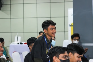 Several young individuals are sitting in a classroom or conference setting. One person is standing and smiling, while others are seated, some wearing masks. The atmosphere appears to be informal and engaging.