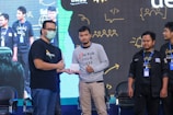 Several individuals posing in a line on a stage, with one person handing an envelope to another. They are wearing casual and branded clothing with identification badges. A large screen in the background displays abstract illustrations and text.
