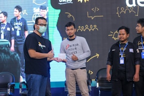 Several individuals posing in a line on a stage, with one person handing an envelope to another. They are wearing casual and branded clothing with identification badges. A large screen in the background displays abstract illustrations and text.