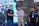 Several individuals posing in a line on a stage, with one person handing an envelope to another. They are wearing casual and branded clothing with identification badges. A large screen in the background displays abstract illustrations and text.