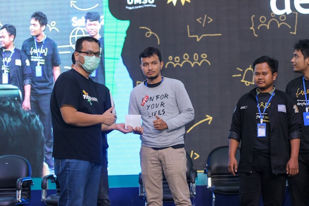 Several individuals posing in a line on a stage, with one person handing an envelope to another. They are wearing casual and branded clothing with identification badges. A large screen in the background displays abstract illustrations and text.