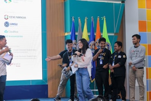A group of people standing on a stage, some wearing lanyards, and holding microphones. The background has multiple colorful flags and a large screen displaying logos and names, indicating the event might be sponsored. The people appear to be in a jovial and engaged state.