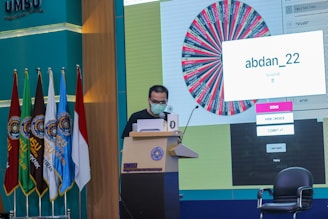 A person stands at a podium speaking into a microphone, wearing a mask and holding papers. Behind them is a large screen displaying a colorful wheel and a message saying 'abdan_22'. Several flags are positioned to the left, and chairs are visible nearby. The setting appears to be a formal event or conference at UMSU.