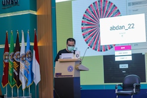 A person stands at a podium speaking into a microphone, wearing a mask and holding papers. Behind them is a large screen displaying a colorful wheel and a message saying 'abdan_22'. Several flags are positioned to the left, and chairs are visible nearby. The setting appears to be a formal event or conference at UMSU.