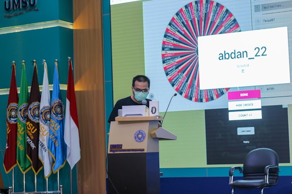 A person stands at a podium speaking into a microphone, wearing a mask and holding papers. Behind them is a large screen displaying a colorful wheel and a message saying 'abdan_22'. Several flags are positioned to the left, and chairs are visible nearby. The setting appears to be a formal event or conference at UMSU.