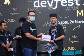 Two individuals stand on a stage, one wearing a mask, handing over a tablet to the other. The background features a large screen displaying event details with the date '26 November'. Other people are present, likely for a tech-related gathering.