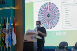 A person wearing a mask stands at a podium in front of a large screen displaying a colorful spinning wheel with names and a list of inputs. Several flags are positioned to the left, and a banner on the podium features UMSU's logo and name. The setting appears to be an indoor conference or seminar.