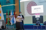 A person is standing at a podium wearing a face mask, participating in an event or presentation. Multiple flags are displayed in a row on the left side, representing an educational or institutional setting. A large screen shows a graphical wheel with text, possibly part of an interactive selection process.