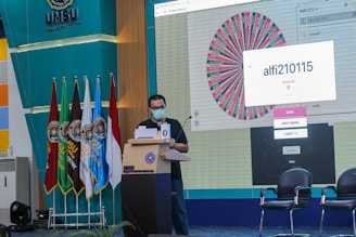 A person is standing at a podium wearing a face mask, participating in an event or presentation. Multiple flags are displayed in a row on the left side, representing an educational or institutional setting. A large screen shows a graphical wheel with text, possibly part of an interactive selection process.