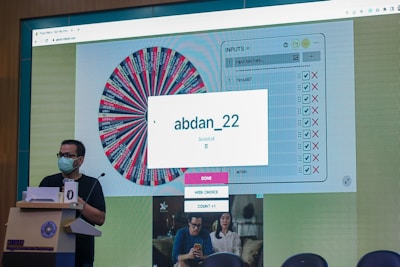 A person wearing a face mask stands at a podium with a microphone. In the background, a large screen displays a digital wheel with colorful segments and various names on it. A box on the screen highlights a selected name 'abdan_22' with a message indicating it is chosen.