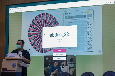 A person wearing a face mask stands at a podium with a microphone. In the background, a large screen displays a digital wheel with colorful segments and various names on it. A box on the screen highlights a selected name 'abdan_22' with a message indicating it is chosen.