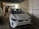 A white car is parked inside a dimly lit garage beside another vehicle. The walls are unfinished and there's a mix of industrial elements surrounding the space.