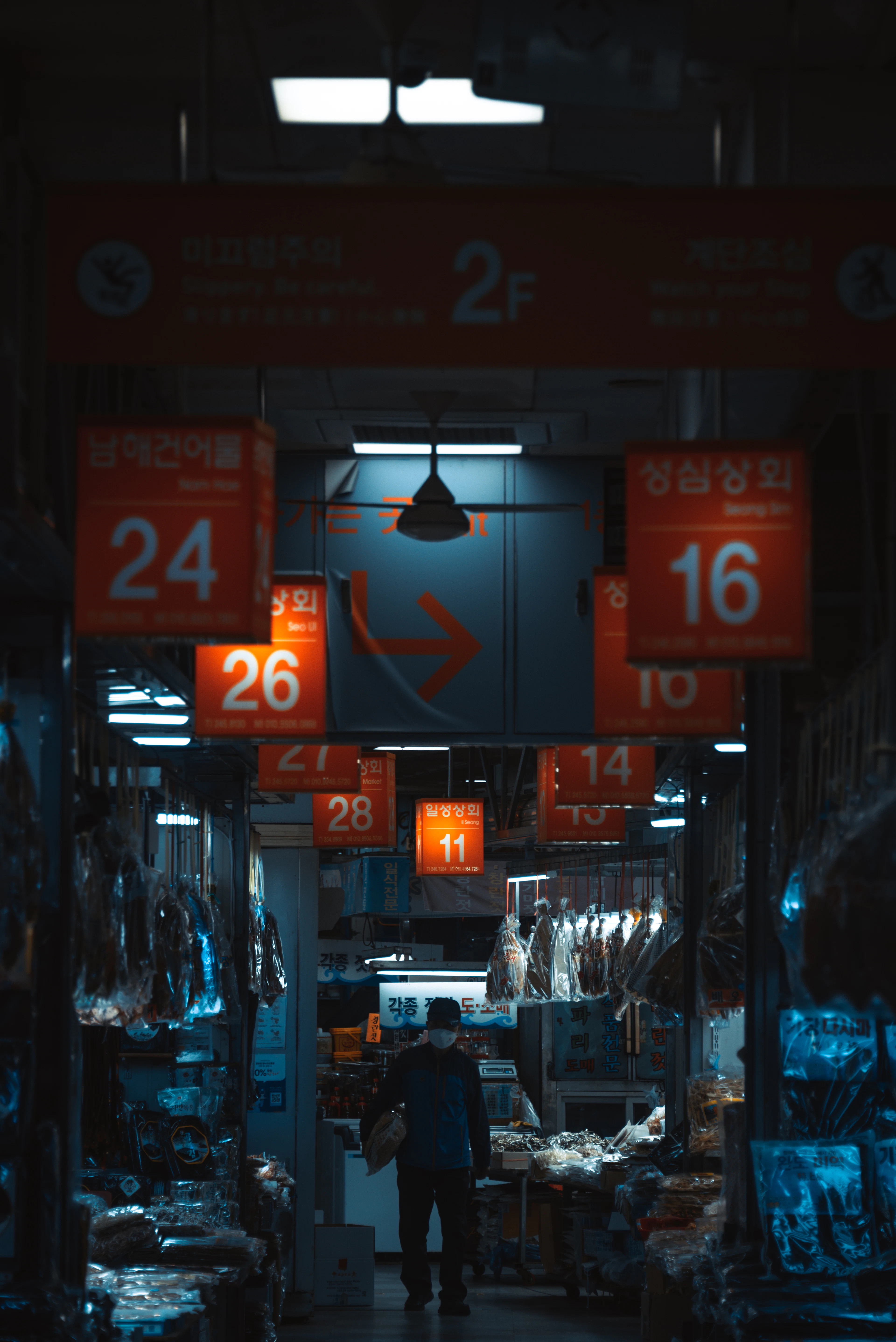 A shopper walks through a dim indoor seafood market in Busan, red numbered stall signs glowing overhead.