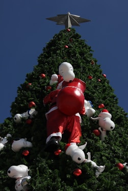 A large Christmas tree is adorned with numerous figures of a cartoon dog, some dressed as Santa Claus. The tree is richly decorated with red ornaments and topped with a silver star. The figures are positioned playfully among the branches.
