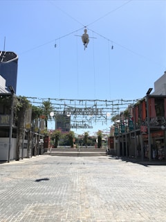 A spacious outdoor plaza with cobblestone paving and storefronts on either side. Greenery is hanging from string lights above the square, and a large decorative ornament or fixture is suspended centrally from wires. The weather is sunny with a clear blue sky. In the background, trees and a building are visible.