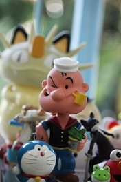 A collection of cartoon character figurines, including a sailor with a pipe and a tin of spinach, a robotic cat with a bell on its collar, a frog, and other colorful toys. They are set against a blurred background with natural light filtering through.