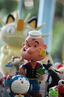 A collection of cartoon character figurines, including a sailor with a pipe and a tin of spinach, a robotic cat with a bell on its collar, a frog, and other colorful toys. They are set against a blurred background with natural light filtering through.