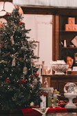 A Christmas tree decorated with various ornaments and lights, placed in a welcoming indoor setting. Underneath the tree, there are wrapped gifts and a decorative snow globe. In the background, a person is seen working at a wooden counter that holds various items including a large decorative bowl and some gift boxes.