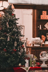 A Christmas tree decorated with various ornaments and lights, placed in a welcoming indoor setting. Underneath the tree, there are wrapped gifts and a decorative snow globe. In the background, a person is seen working at a wooden counter that holds various items including a large decorative bowl and some gift boxes.