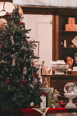 A Christmas tree decorated with various ornaments and lights, placed in a welcoming indoor setting. Underneath the tree, there are wrapped gifts and a decorative snow globe. In the background, a person is seen working at a wooden counter that holds various items including a large decorative bowl and some gift boxes.