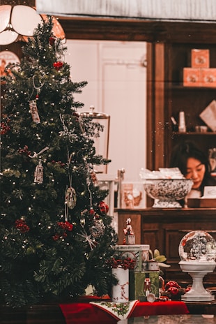 A Christmas tree decorated with various ornaments and lights, placed in a welcoming indoor setting. Underneath the tree, there are wrapped gifts and a decorative snow globe. In the background, a person is seen working at a wooden counter that holds various items including a large decorative bowl and some gift boxes.
