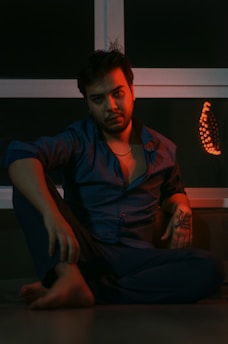 A seated individual with a contemplative expression is positioned against a window backdrop. Warm red and orange lighting casts a dramatic shadow, highlighting the person's facial features and the tattoo on their arm. The overall setting seems casual yet introspective, with the person wearing an open dress shirt and pants.