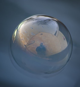 A reflective surface captures the image of a person standing in a dark coat. The scene appears to be enclosed in a spherical, transparent medium with a glossy finish, exhibiting a clear reflection of the surroundings. The environment includes muted colors and subtle lighting that enhances the swirls and texture on the surface.