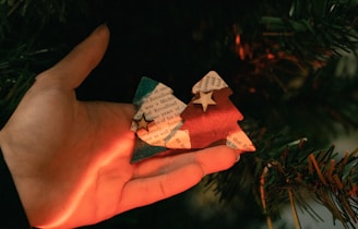 A hand holds two small tree-shaped ornaments with a rustic design, made from paper. One tree is red, and the other is green, both featuring star embellishments on top. Soft, warm lighting casts a gentle glow on the hand and ornaments, creating a cozy ambiance. The background includes blurred green foliage, resembling a Christmas tree.