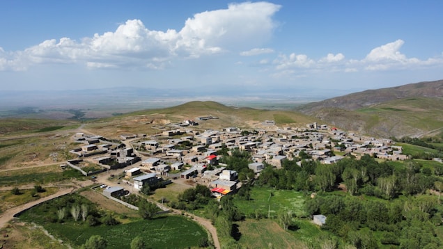 A panoramic view of a small village situated in a hilly landscape. The village consists of numerous rectangular buildings made from light-colored materials, scattered across the green terrain. Lush greenery surrounds the structures, and there is a mix of open fields and trees. The sky is mostly clear with a few scattered clouds, creating a tranquil rural setting.
