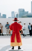 A person dressed in traditional attire stands facing a group of people. The attire includes a red robe with yellow trim, a hat adorned with feathers, and decorative wrist coverings. Several individuals in casual clothing are in the background, with tall buildings also visible, suggesting an urban setting.