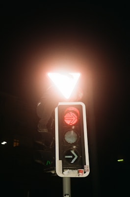 A traffic light against a dark background displays a red illuminated arrow pointing to the right. Above the traffic light is a triangular road sign that glows brightly with white light.
