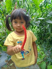 A young child with dark hair is standing among lush green chili plants. The child is wearing a yellow shirt with a cartoon character on it and is holding a long red chili pepper in one hand, extending it towards the camera. The surrounding environment is rich with green leaves and soil is visible on the ground.