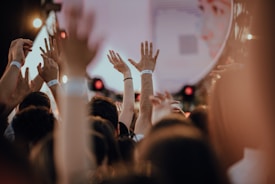 A crowd of people with their hands raised, suggesting excitement and engagement. Wristbands are visible, indicating attendees of an event, such as a concert or festival. The background includes blurred lights and what appears to be a stage setup.
