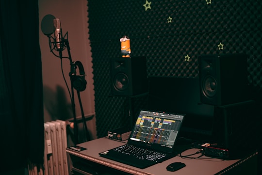A dark, cozy music studio with a microphone and headphones on a stand to the left, positioned in front of sound-absorbing foam panels. On the desk, a laptop with an audio mixing application open is flanked by two studio monitor speakers. Small, star-shaped decorations are scattered on the foam wall, adding a whimsical touch. A small figurine sits atop one of the speakers.