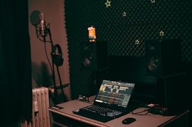 A dark, cozy music studio with a microphone and headphones on a stand to the left, positioned in front of sound-absorbing foam panels. On the desk, a laptop with an audio mixing application open is flanked by two studio monitor speakers. Small, star-shaped decorations are scattered on the foam wall, adding a whimsical touch. A small figurine sits atop one of the speakers.