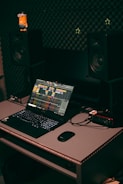 A home studio setup featuring a laptop with music production software open, set on a desk with a mouse and audio interface. Two large studio monitors are positioned on either side, with soundproofing material on the wall behind them. A small toy figure is placed on one of the monitors.