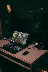 A home studio setup featuring a laptop with music production software open, set on a desk with a mouse and audio interface. Two large studio monitors are positioned on either side, with soundproofing material on the wall behind them. A small toy figure is placed on one of the monitors.
