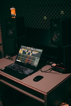 A home studio setup featuring a laptop with music production software open, set on a desk with a mouse and audio interface. Two large studio monitors are positioned on either side, with soundproofing material on the wall behind them. A small toy figure is placed on one of the monitors.