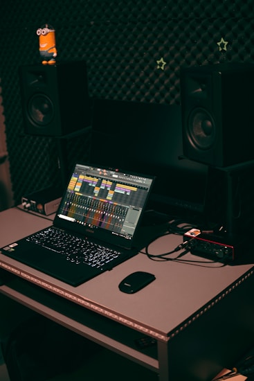 A home studio setup featuring a laptop with music production software open, set on a desk with a mouse and audio interface. Two large studio monitors are positioned on either side, with soundproofing material on the wall behind them. A small toy figure is placed on one of the monitors.
