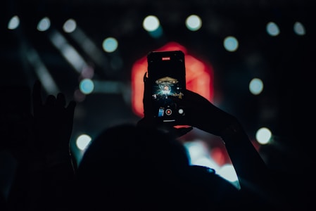 A silhouetted hand holds up a smartphone to record a concert or event, with a blurred stage and bright lights in the background.