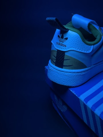 A close-up view of the back of an Adidas sneaker resting on a shoebox under a blue light. The sneaker has a leather texture, with a visible brand logo and a contrast between green and white colors.