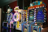 A row of brightly colored arcade game machines is arranged in a line against the wall. Each machine features vibrant lights and designs, with themes ranging from claw games to game show-inspired setups. The area has a playful and inviting atmosphere, suitable for entertainment or amusement centers.