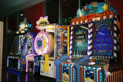 A row of brightly colored arcade game machines is arranged in a line against the wall. Each machine features vibrant lights and designs, with themes ranging from claw games to game show-inspired setups. The area has a playful and inviting atmosphere, suitable for entertainment or amusement centers.