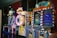 A row of brightly colored arcade game machines is arranged in a line against the wall. Each machine features vibrant lights and designs, with themes ranging from claw games to game show-inspired setups. The area has a playful and inviting atmosphere, suitable for entertainment or amusement centers.