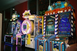 A row of brightly colored arcade game machines is arranged in a line against the wall. Each machine features vibrant lights and designs, with themes ranging from claw games to game show-inspired setups. The area has a playful and inviting atmosphere, suitable for entertainment or amusement centers.