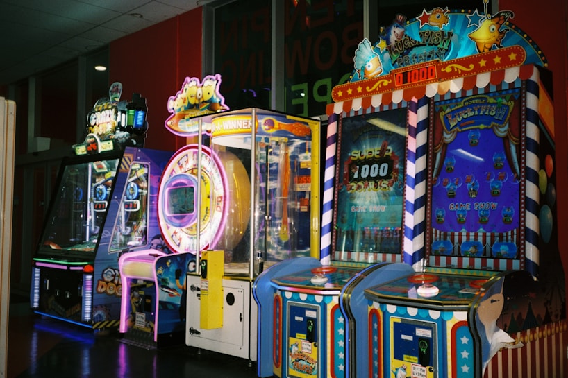 A row of brightly colored arcade game machines is arranged in a line against the wall. Each machine features vibrant lights and designs, with themes ranging from claw games to game show-inspired setups. The area has a playful and inviting atmosphere, suitable for entertainment or amusement centers.