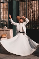A man dressed in traditional white clothing and a tall, brown hat is performing a dance with one arm raised and the other hand near his head, spinning gracefully. A ginger cat lies on a stone ledge next to him, basking in the sunlight. The background features a wrought iron fence and trees with bare branches, suggesting an outdoor urban setting.