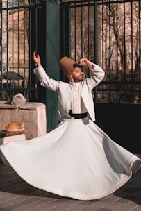 A man dressed in traditional white clothing and a tall, brown hat is performing a dance with one arm raised and the other hand near his head, spinning gracefully. A ginger cat lies on a stone ledge next to him, basking in the sunlight. The background features a wrought iron fence and trees with bare branches, suggesting an outdoor urban setting.