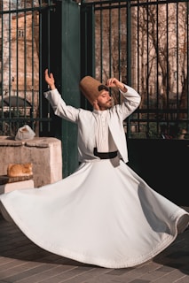 A man dressed in traditional white clothing and a tall, brown hat is performing a dance with one arm raised and the other hand near his head, spinning gracefully. A ginger cat lies on a stone ledge next to him, basking in the sunlight. The background features a wrought iron fence and trees with bare branches, suggesting an outdoor urban setting.
