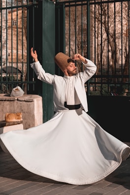 A man dressed in traditional white clothing and a tall, brown hat is performing a dance with one arm raised and the other hand near his head, spinning gracefully. A ginger cat lies on a stone ledge next to him, basking in the sunlight. The background features a wrought iron fence and trees with bare branches, suggesting an outdoor urban setting.